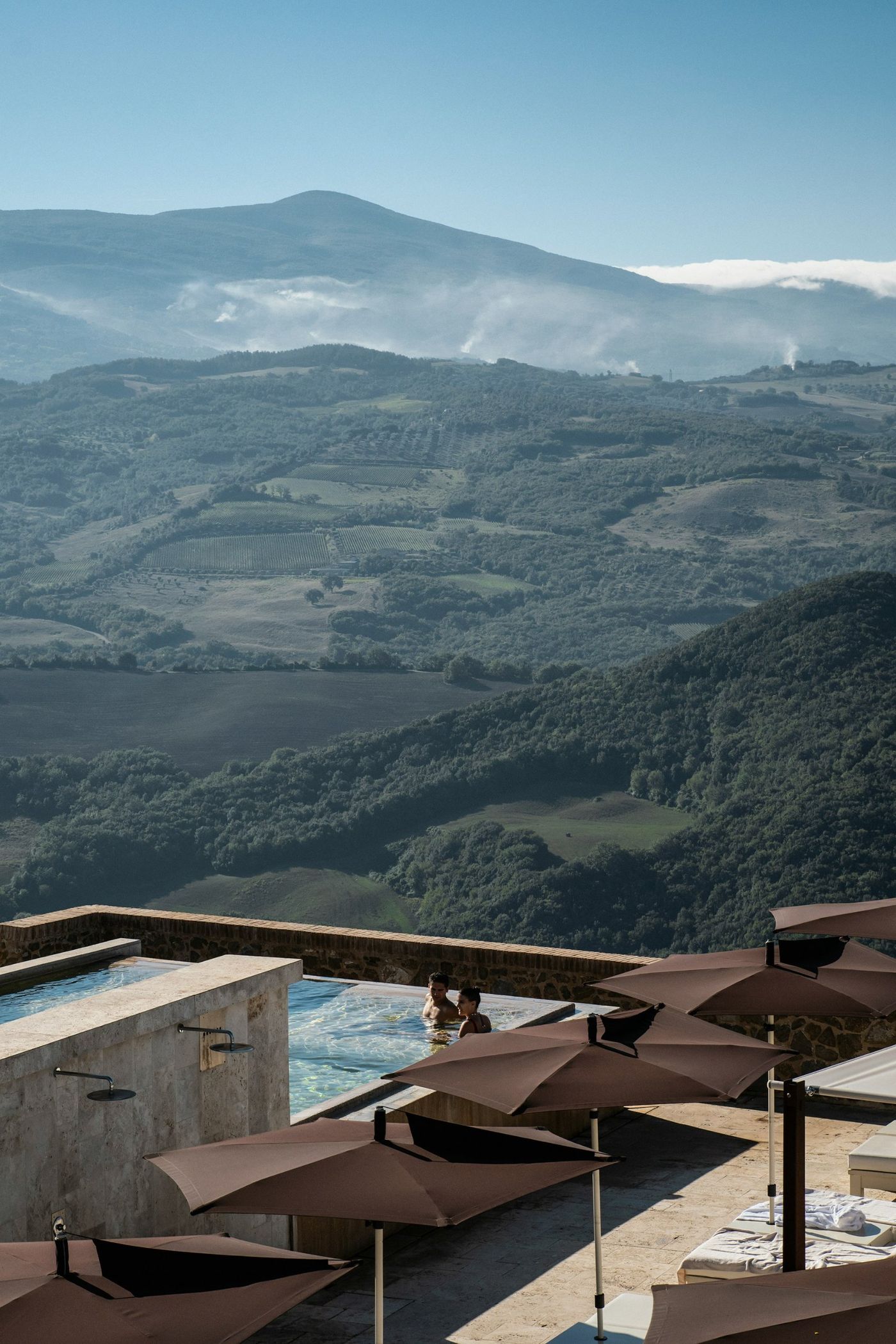 A couple in a stone infinity pool above rolling green hills and vineyards, brown parasols in the foreground