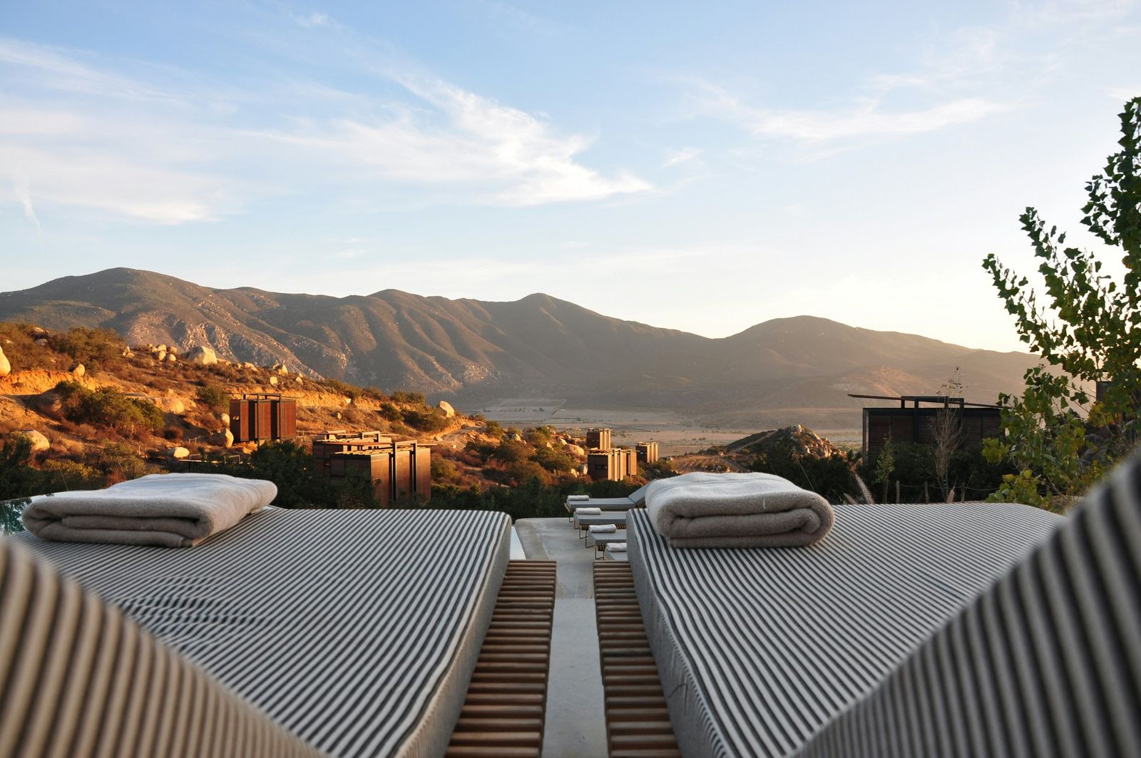 Two striped sunbeds with folded towels facing a mountain valley and scattered cabins at golden hour