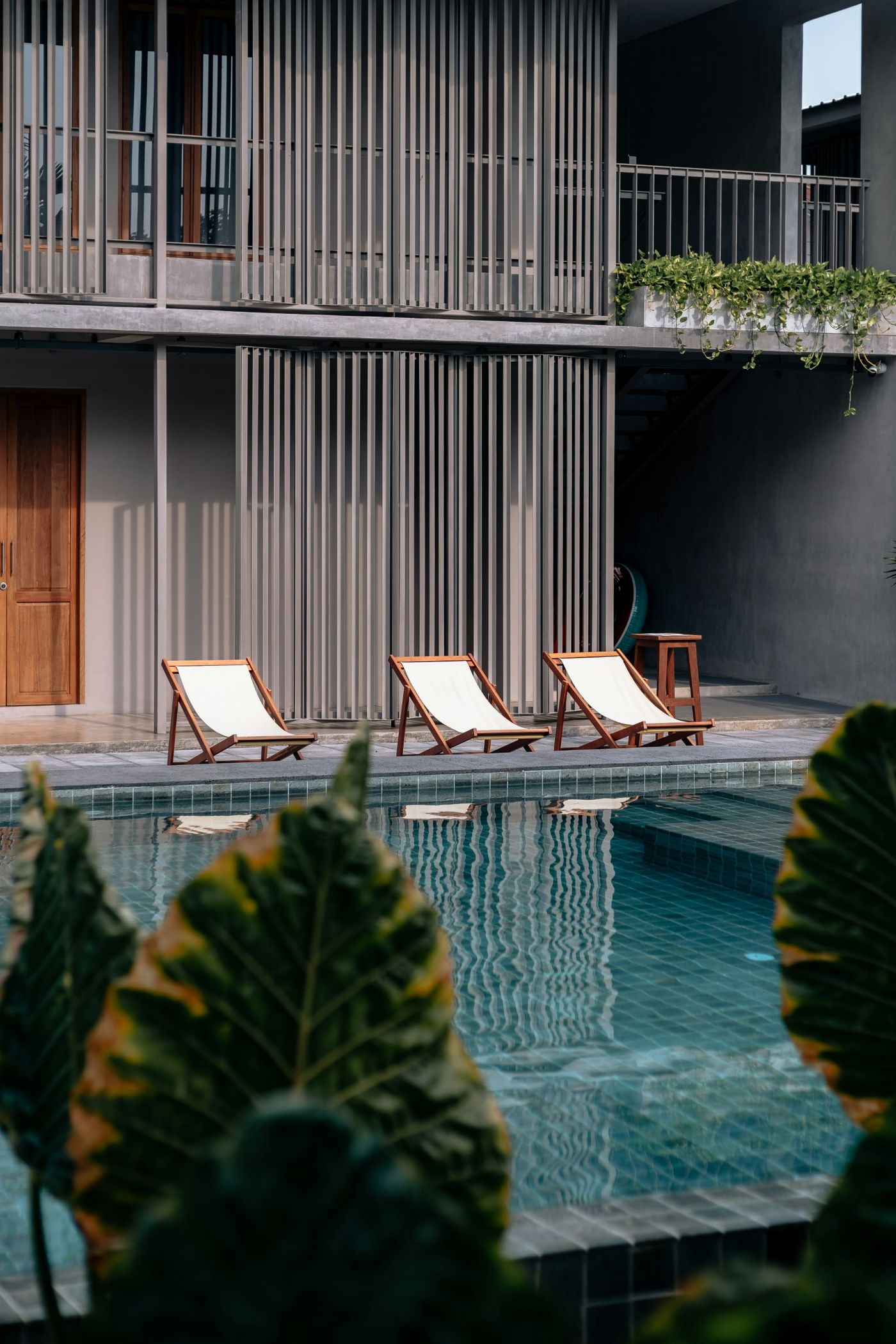Three canvas deck chairs beside a tiled pool in front of a grey facade with vertical wooden screens
