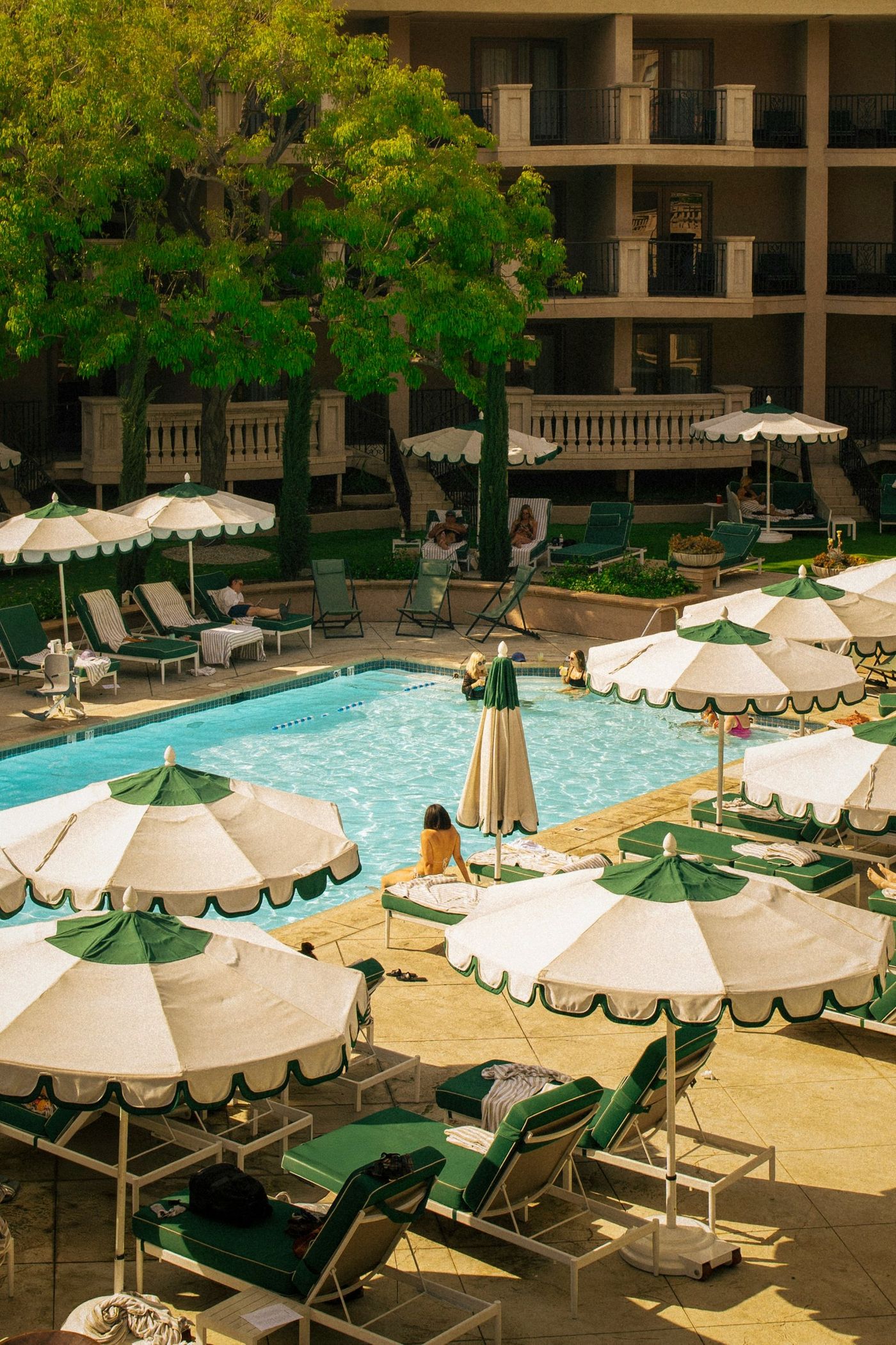 A sunlit hotel pool courtyard with green and white scalloped parasols and loungers