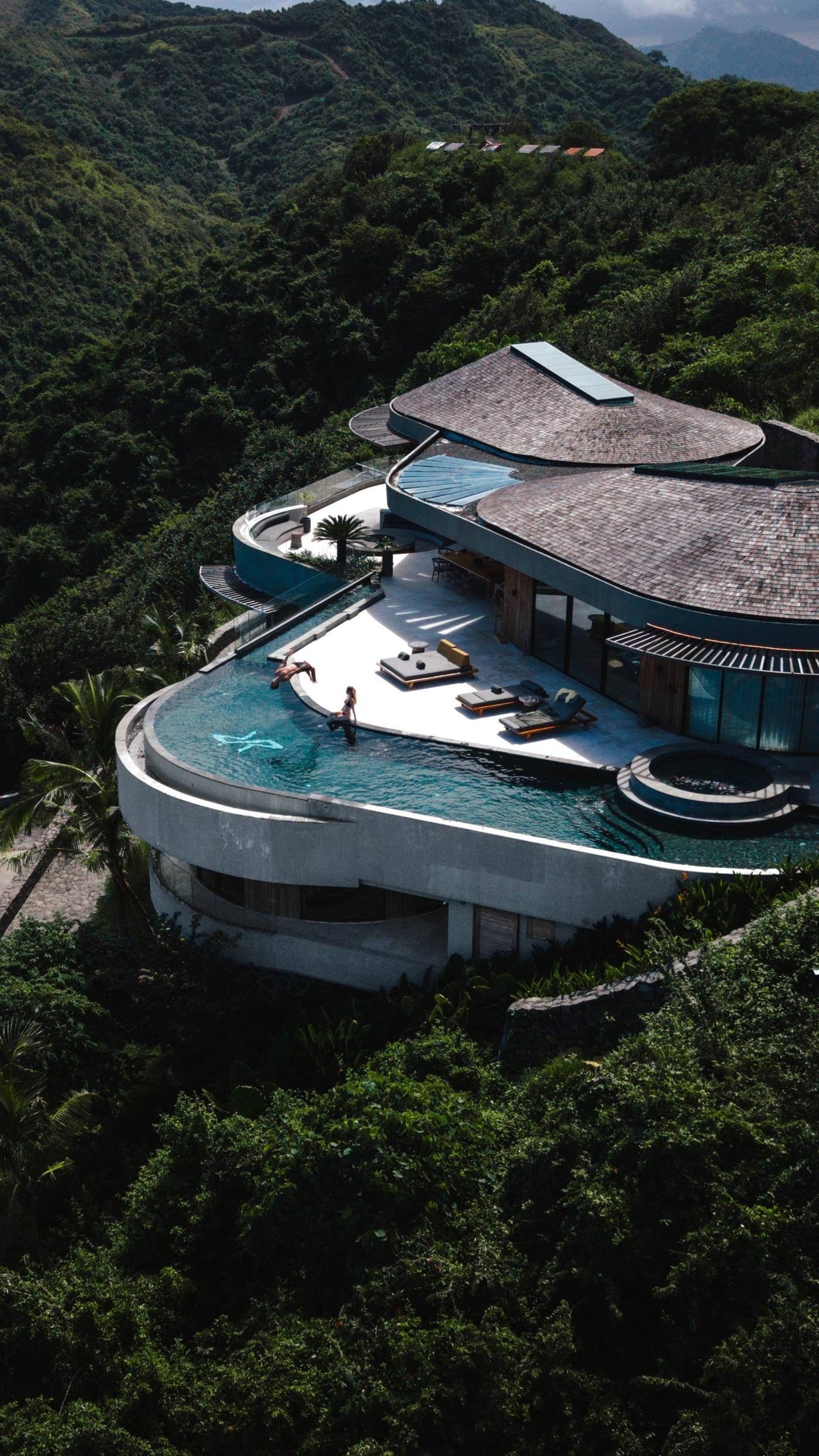 Aerial view of a curved hillside villa with a sweeping infinity pool, surrounded by dense green forest